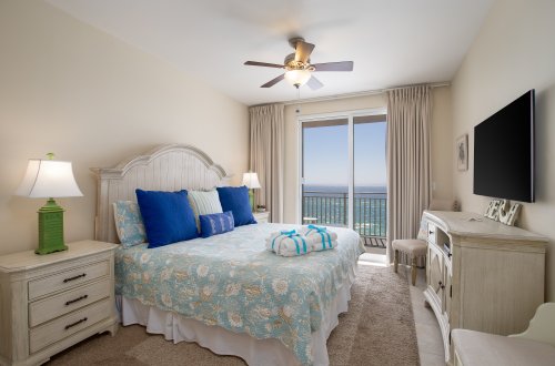 A wide view of a guest room with a bed, dresser, TV, and balcony access with a beach view.