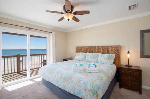 A wide view of a guest room with a bed, nightstand, ceiling fan, and sliding doors leading to a balcony with a view of the ocean.