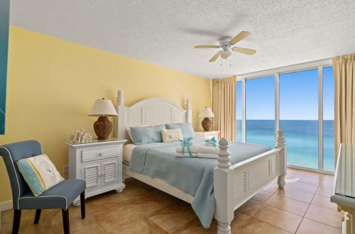 A wide view of a hotel room with a bed, chair, nightstand, and large windows showing an ocean view.