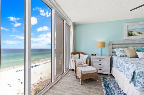 A beachfront hotel room with a large window showing an ocean view, a bed, chair, and nightstand.