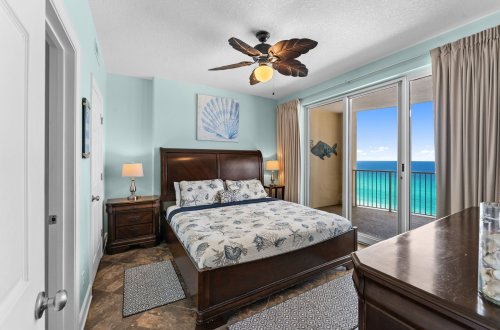 A wide view of a bedroom with a large bed, nightstands, a ceiling fan, and a sliding glass door leading to a balcony with an ocean view.