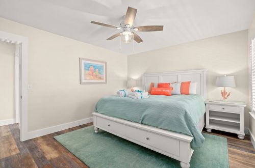 A neatly made bed with a teal comforter and orange pillows in a room with a ceiling fan and a nightstand.