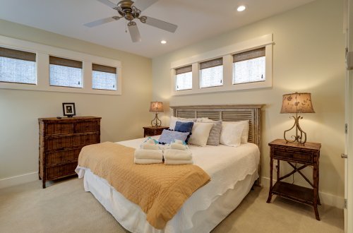 A neatly made bed with a white comforter and pillows, a wooden dresser, and two nightstands with lamps in a beige-painted bedroom with windows and a ceiling fan.
