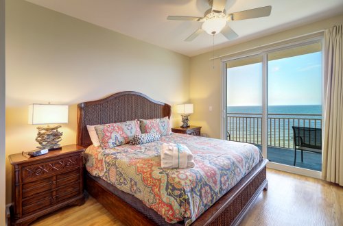 A wide-angle view of a guest room with a bed, nightstands, lamps, and a ceiling fan, featuring a large window with a ocean view.