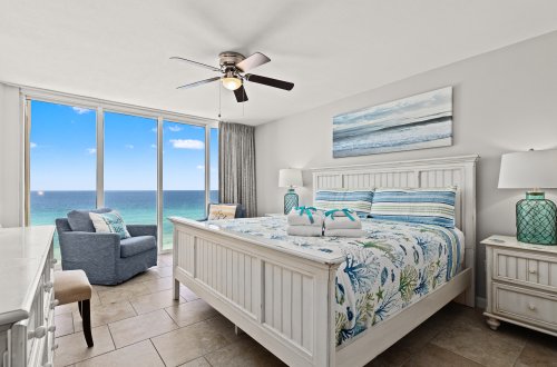 A wide view of a beach-themed guest room with a large bed, nightstands, a chair, and a ceiling fan, featuring a large window with a view of the ocean.