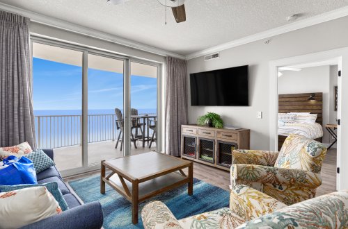 A wide-angle view of a living room with a balcony overlooking the ocean, a dining area, and a bedroom in the background.