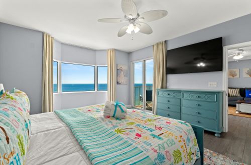 A wide-angle view of a beach-themed bedroom with a large bed, dresser, and TV, featuring a stunning ocean view through the windows.