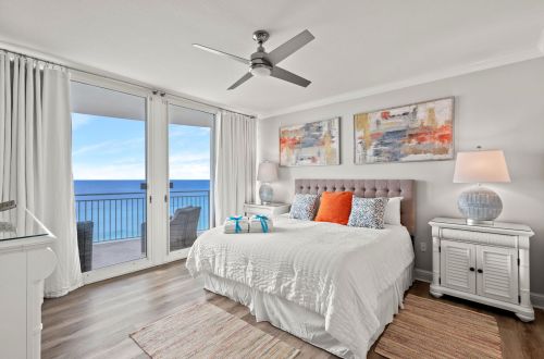 A white bedroom with a large bed, ceiling fan, and balcony with ocean view.