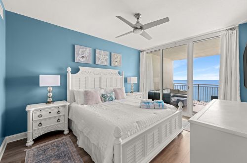 A wide view of a guest room with a white bed, blue wall, ceiling fan, and sliding glass doors leading to a balcony with an ocean view.