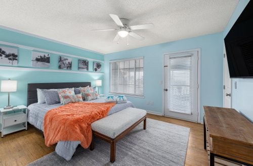 A wide view of a bedroom with a bed, nightstand, lamp, TV, ceiling fan, window, and door.