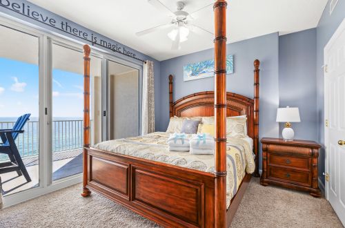 A wide view of a guest room with a four-poster bed, nightstand, and sliding glass door leading to a balcony with an ocean view.