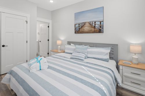 A neatly made bed with a blue and white striped comforter, two nightstands with lamps, and a painting of a dock above the bed.