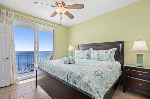 A wide view of a bedroom with a large bed, nightstands, lamps, and a sliding glass door leading to a balcony with a view of the ocean.