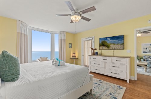 A wide view of a bright guest room with a large bed, ocean view, and white furniture.