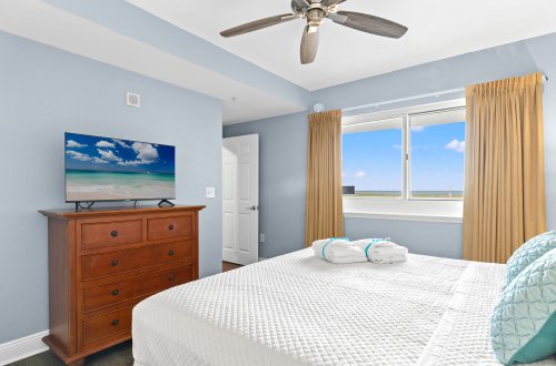 A bedroom with a large bed, TV on a dresser, window with a view, and ceiling fan.