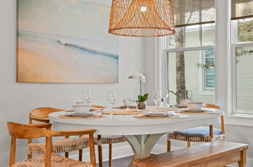 A dining area with a round table set for dinner, a woven light fixture, a beach-themed painting, and a window with a view.
