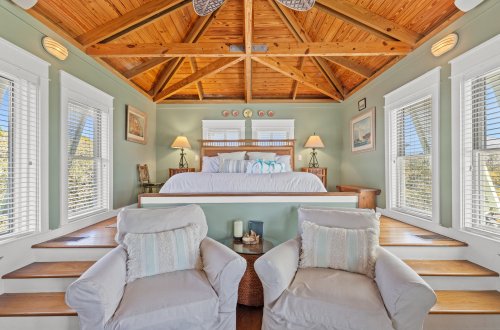 A spacious room with a vaulted ceiling, wooden beams, and large windows, featuring a raised bed platform and seating area.