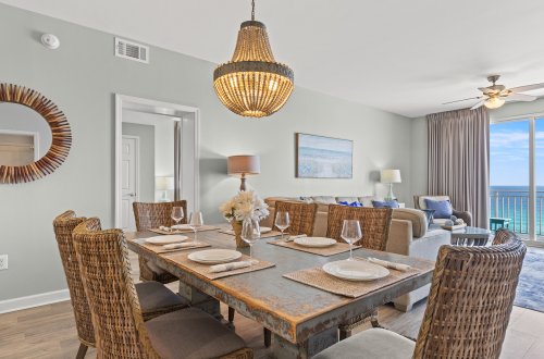 A dining area with a wooden table, wicker chairs, and a chandelier, adjacent to a living area with a couch and a large window.
