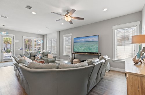 A bright and airy living room with a large gray sectional sofa, a TV mounted on the wall, and a ceiling fan.