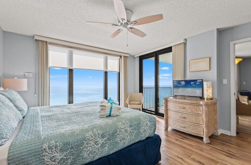 A wide view of a bedroom with a bed, dresser, ceiling fan, and large window showing a ocean view.